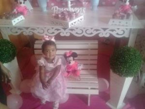 Foto de menina de um ano sentada em banco branco, vestida de cor-de-rosa
