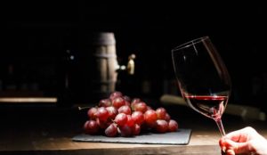 Foto de um pequeno tonel de vinho, um cacho de uva e uma mão segurando uma taça com um pouco de vinho tinto