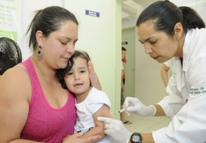 Criança com menos de seis anos recebe dose de vacina sentada no colo de mulher