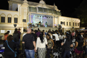 Foto noturna do palco do Coreto iluminado, com apresentação de banda e público assistindo sentado e em pé